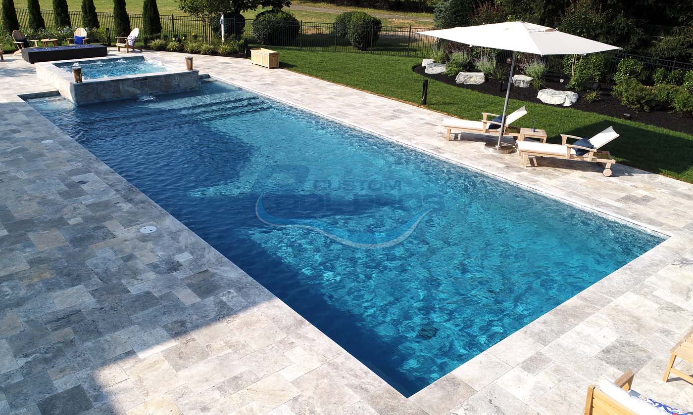 Aerial view of a luxurious outdoor swimming pool with clear blue water, surrounded by elegant stone decking, lounge chairs, and a shaded area with an umbrella, set in a lush green garden.
