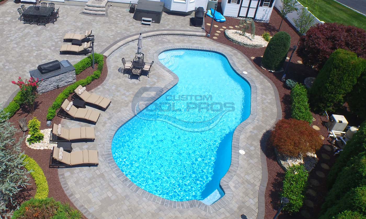 Aerial view of a beautifully designed residential swimming pool surrounded by lounge chairs and landscaped greenery, showcasing a clear blue water surface, pool builder nj