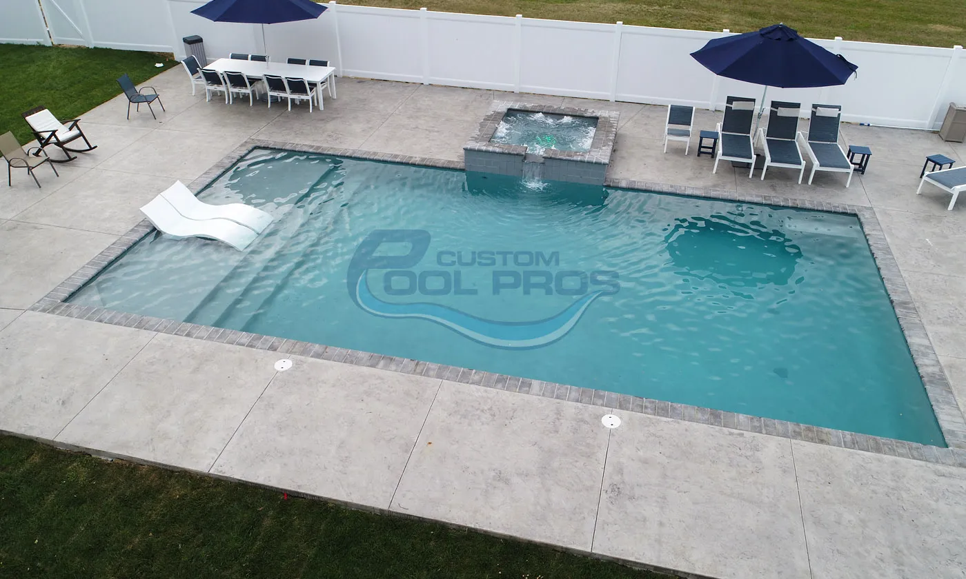 Aerial view of a modern backyard pool with a spa, surrounded by a concrete patio, lounge chairs, and a dining area under umbrellas, showcasing a clean and inviting outdoor space.