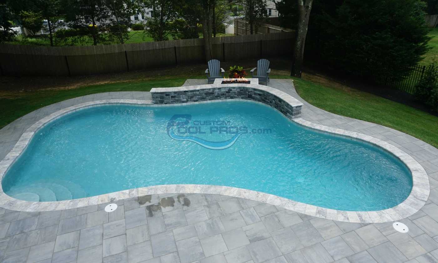 Aerial view of a modern backyard swimming pool with a curved design, surrounded by a stone patio and lush green landscaping. The pool features clear blue water and a built-in seating area with stone accents.