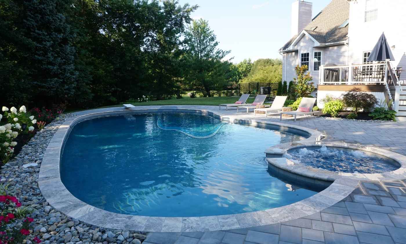 Residential backyard pool with a curved design, surrounded by lush greenery and lounge chairs, featuring a spa area and a nearby house, ideal for relaxation and outdoor gatherings, custom pool builder