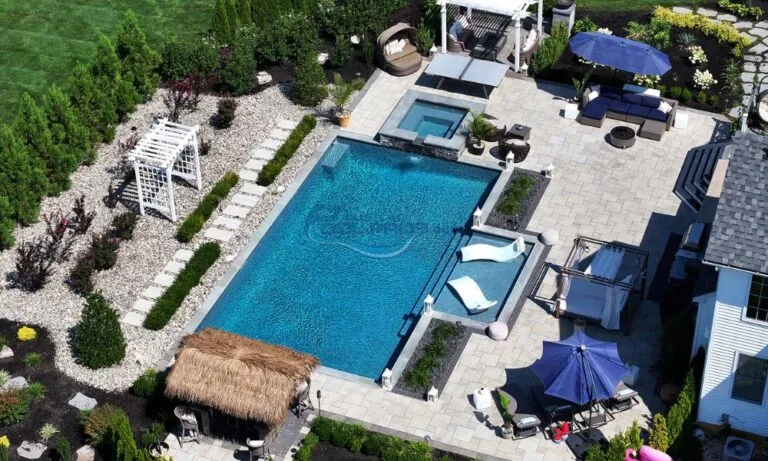 Aerial view of a luxurious swimming pool surrounded by lounge chairs, umbrellas, and landscaped gardens, featuring a hot tub and a tiki bar area for relaxation, luxury swimming pools