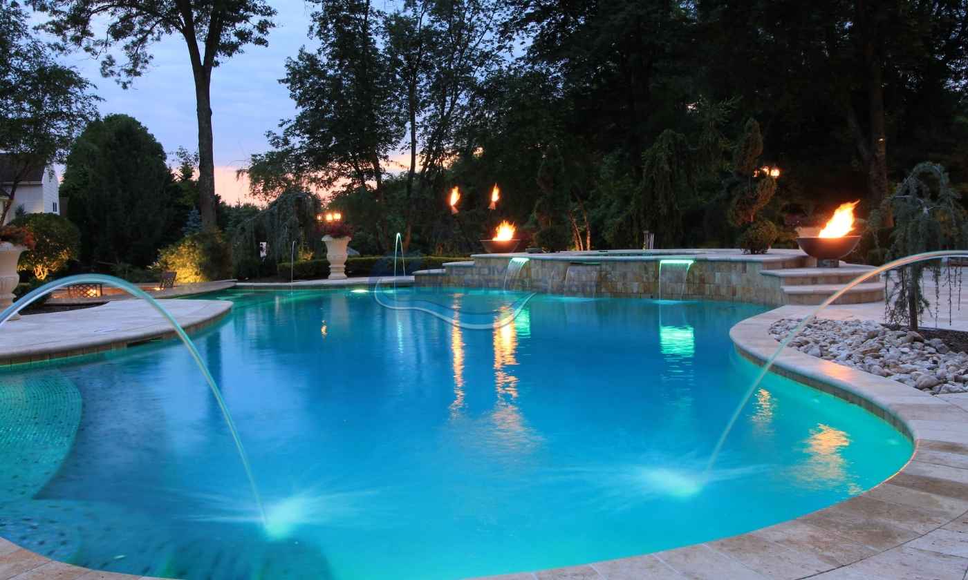 Serene outdoor swimming pool illuminated at dusk, surrounded by lush trees and soft landscape lighting, creating a tranquil atmosphere.