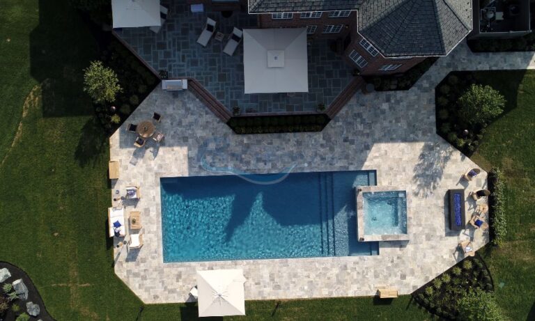 Aerial view of a luxurious backyard featuring a large swimming pool, surrounded by stone patio, lounge chairs, and shaded cabanas, with a well-maintained lawn in a residential setting, pool builder, inground pool contractors